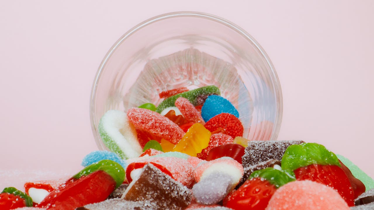 Vibrant sweet gummies spilling from a bowl on a pastel background.